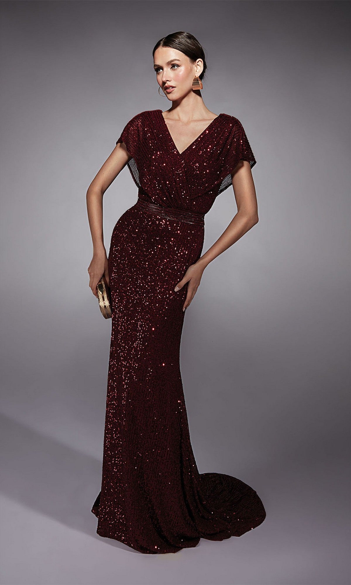 A woman poses against a gray background in the Alyce 27708 long formal dress in wine, featuring sequins, short sleeves, a beaded waistband, and holding a small clutch.